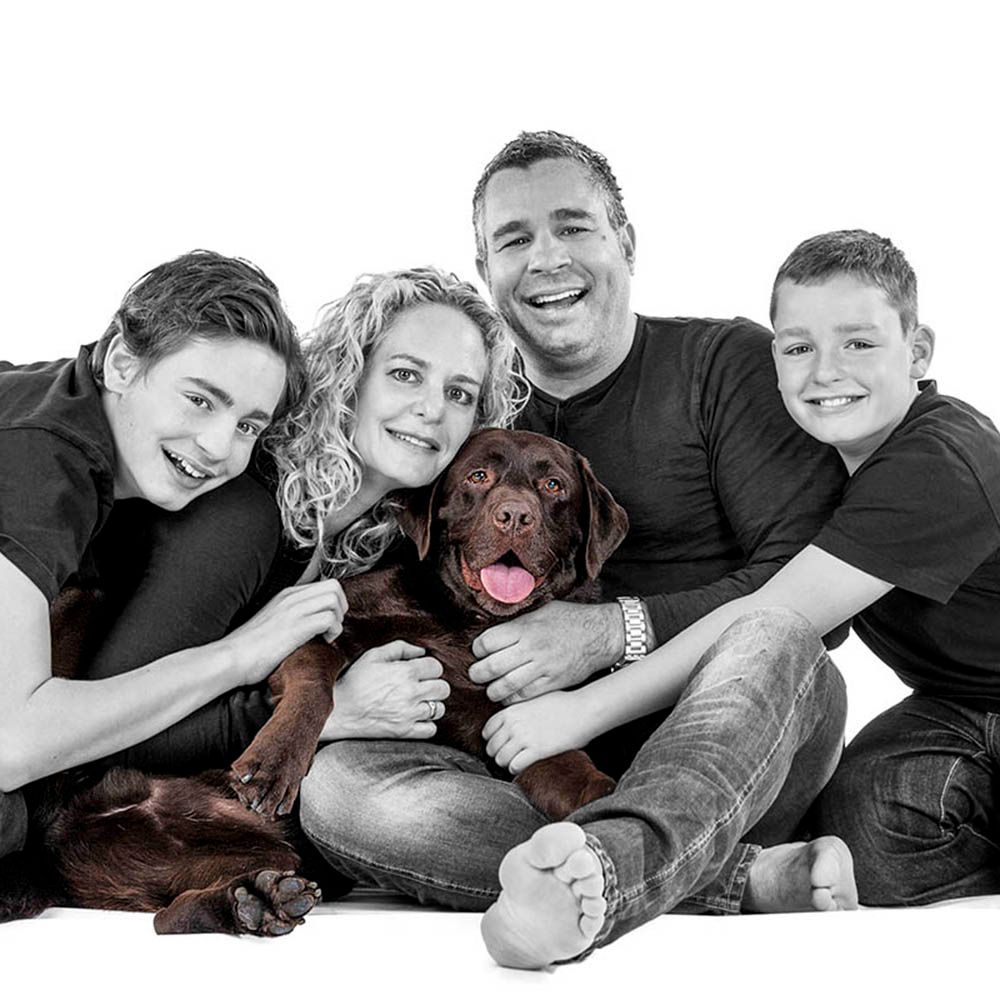 A family of four laughing on the floor with their chocolate labrador