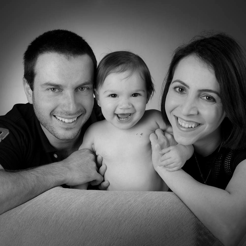 A mother and father cheek to cheek with their laughing baby, in black and white